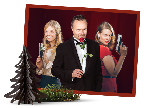 Drei Personen posieren auf einem gerahmten Foto: Ein Mann im Smoking, der eine Rose im Mund hält, steht zwischen zwei Frauen, die mit Spielzeugpistolen zielen. Im Vordergrund befinden sich ein kleiner dekorativer Baum und Grünpflanzen.