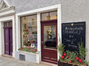 Ein Schokoladengeschäft mit einem geschmückten Weihnachtsbaum im Schaufenster, festlichen roten Blumen draußen und einem Kreideschild in deutscher Sprache, das für handgemachte Schokolade wirbt. Das Schaufenster hat bordeauxfarbene Türen und cremefarbene Verzierungen.