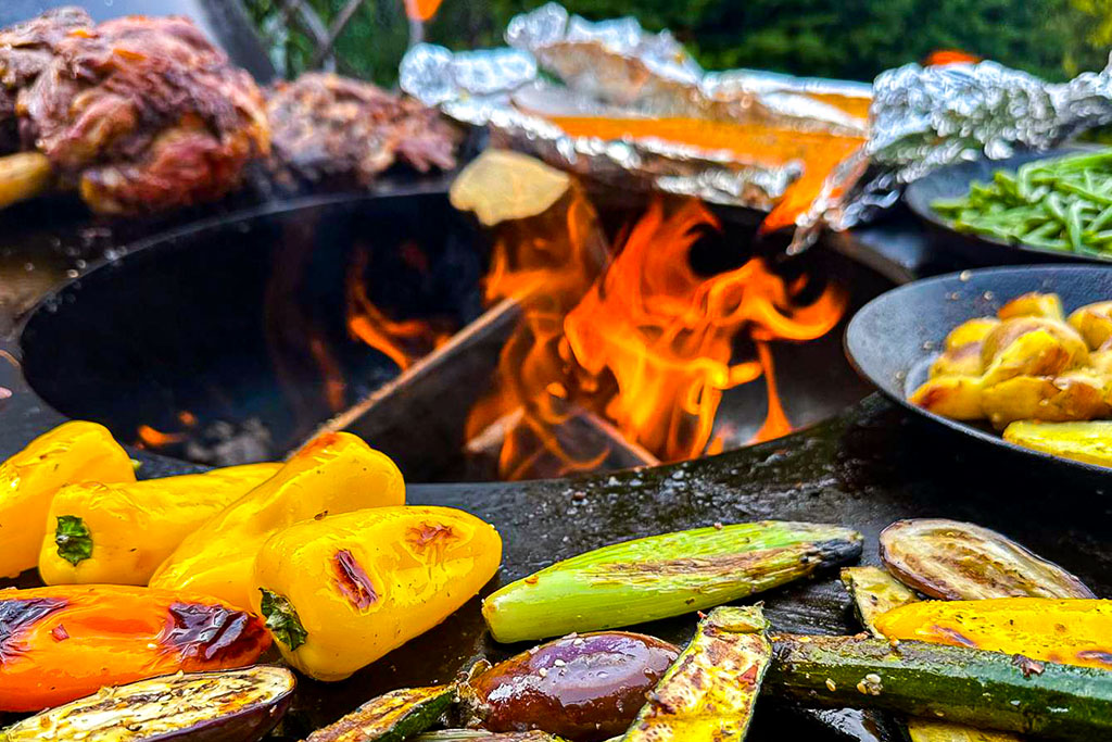 Buntes Gemüse und Fleisch werden über einer offenen Flamme gegrillt, wobei einige Lebensmittel in Folie eingewickelt sind. Leuchtend gelbe Paprika-, Zucchini- und Auberginenscheiben sind im Vordergrund neben dem Feuer zu sehen.