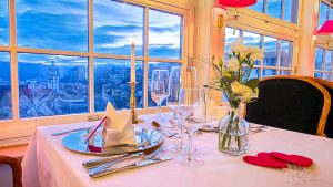 Ein romantisch gedeckter Tisch für zwei Personen an einem großen Fenster, mit gefalteten Servietten, Gläsern, Besteck, einer Vase mit weißen Blumen, roten Herz-Dekorationen und einem Blick auf den Sonnenuntergang über einer Stadt.