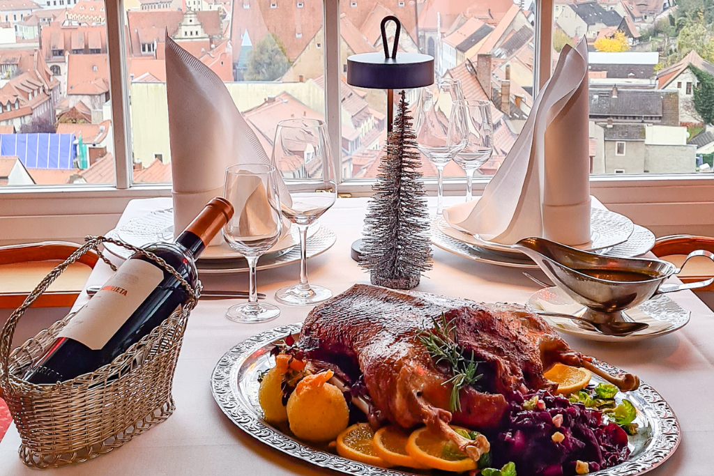 Am Fenster ist ein festlich gedeckter Tisch zu sehen, auf dem gebratene Ente mit Zitrusfrüchten, Wein in einem Korb, Gläser, eine Soßenschüssel und ein kleiner silberner Weihnachtsbaum stehen, während draußen eine Stadtlandschaft zu sehen ist.